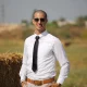 Well-dressed man in sunglasses smiling in a sunlit field, exuding elegance.