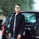 Young man in denim jacket posing confidently with a black SUV in an outdoor setting. Ideal for lifestyle and fashion concepts.