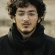 Close-up portrait of a young man with curly hair wearing glasses and a scarf outdoors.